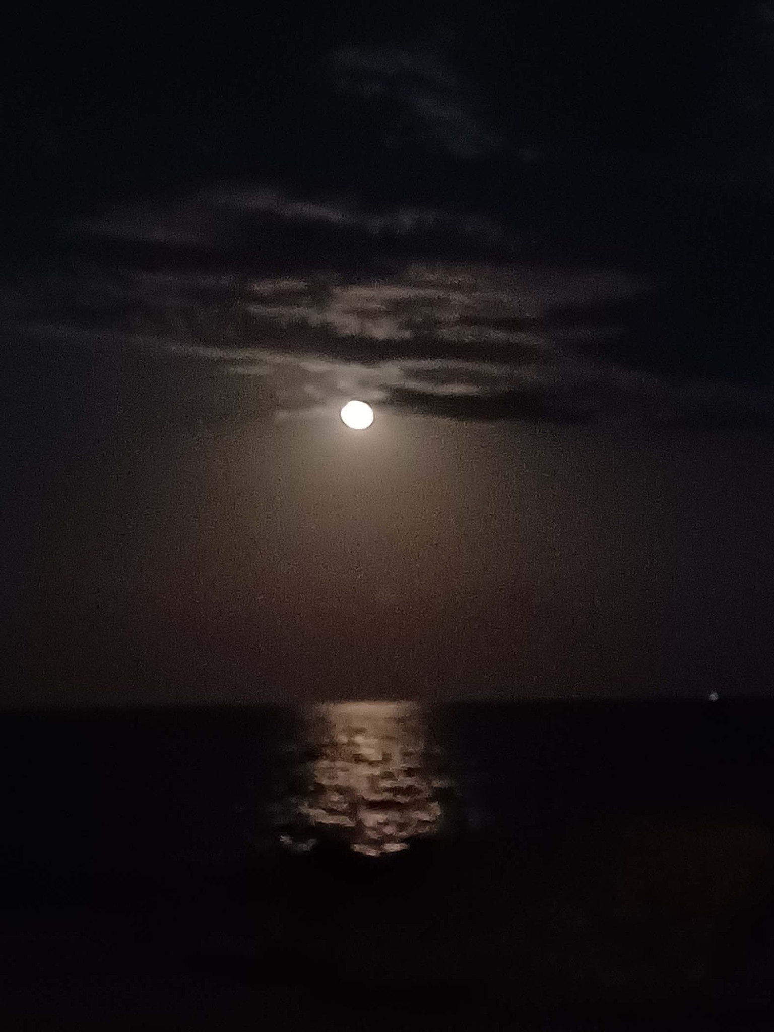 A full moon shining through clouds and reflecting across dark water